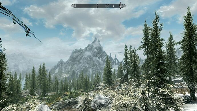 A vista in The Elder Scrolls V: Skyrim showing a blue cloudy sky, pine forest in the foreground, and a snowy mountain in the distance